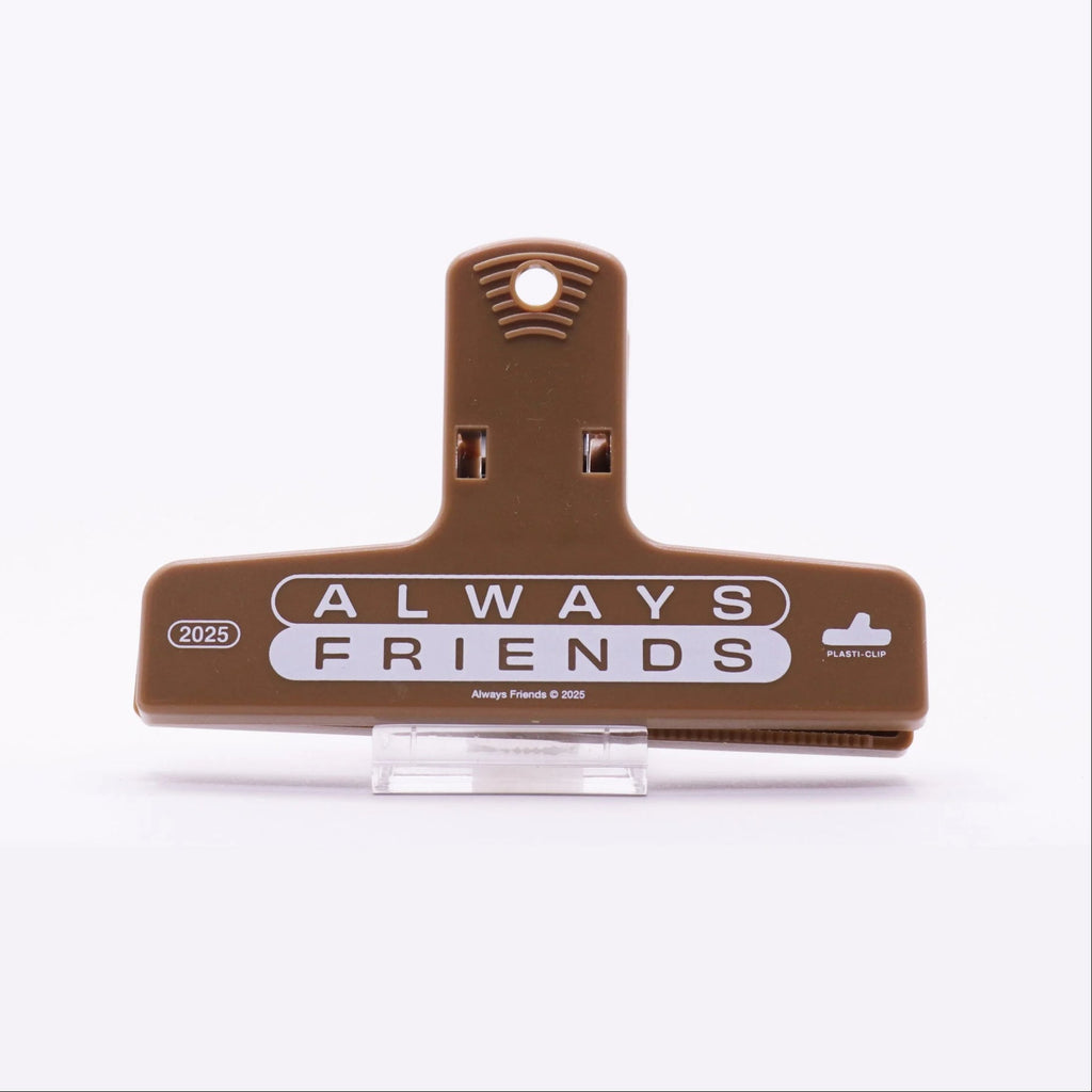 A brown plastic binder clip with black printed text reading 'ALWAYS FRIENDS' and '2025' on the front, displayed on a clear stand against a white background.