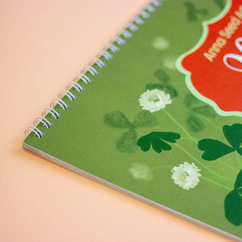 A close-up of a spiral-bound calendar with a green cover featuring shamrock and clover illustrations, white flowers, and a red label area, photographed on a peach background.