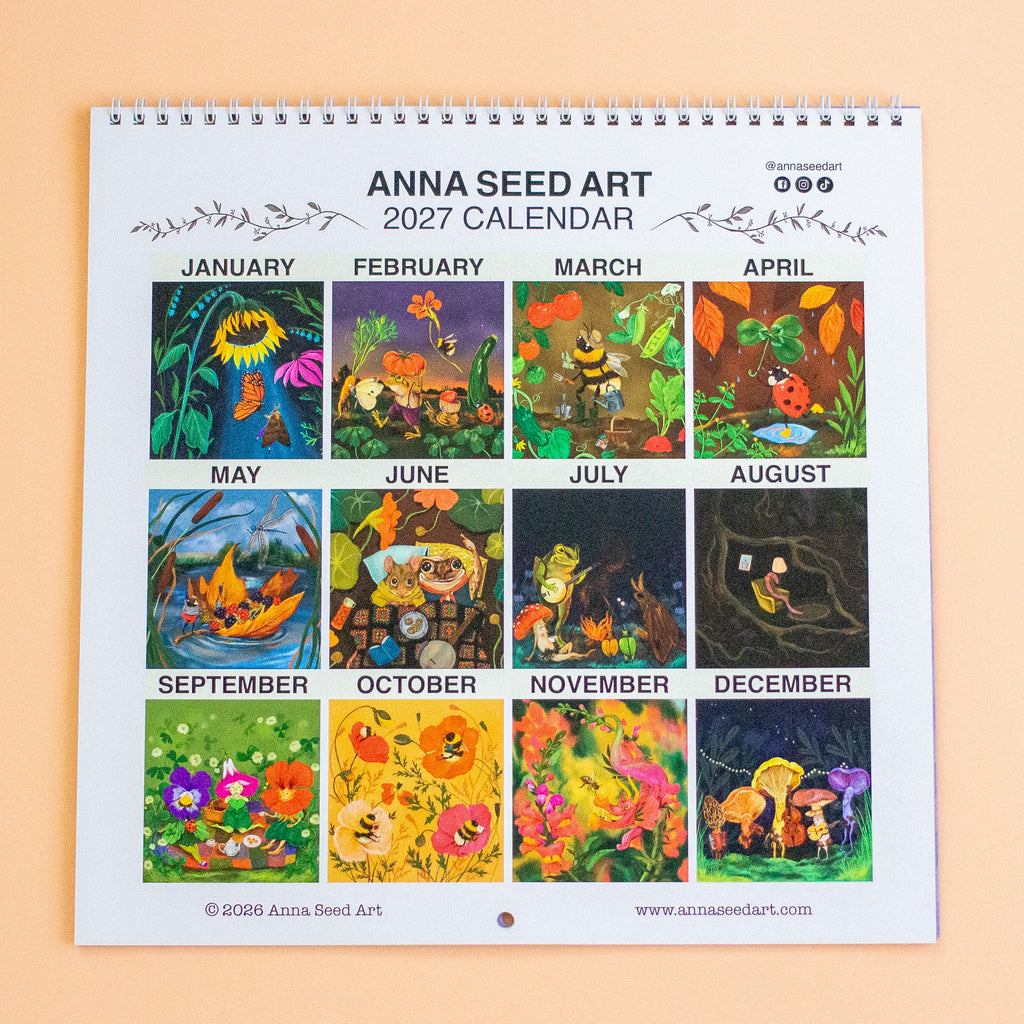 A 2027 spiral-bound Anna Seed Art calendar displayed flat against a peach background. The calendar features 12 square monthly panels with colorful fantasy illustrations, including flowers, mushrooms, and woodland scenes, with labels for each month.