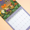 A spiral-bound February wall calendar page with a colourful illustrated cover showing a frog, pumpkin, butterfly, and other garden elements, photographed on a pale peach background.