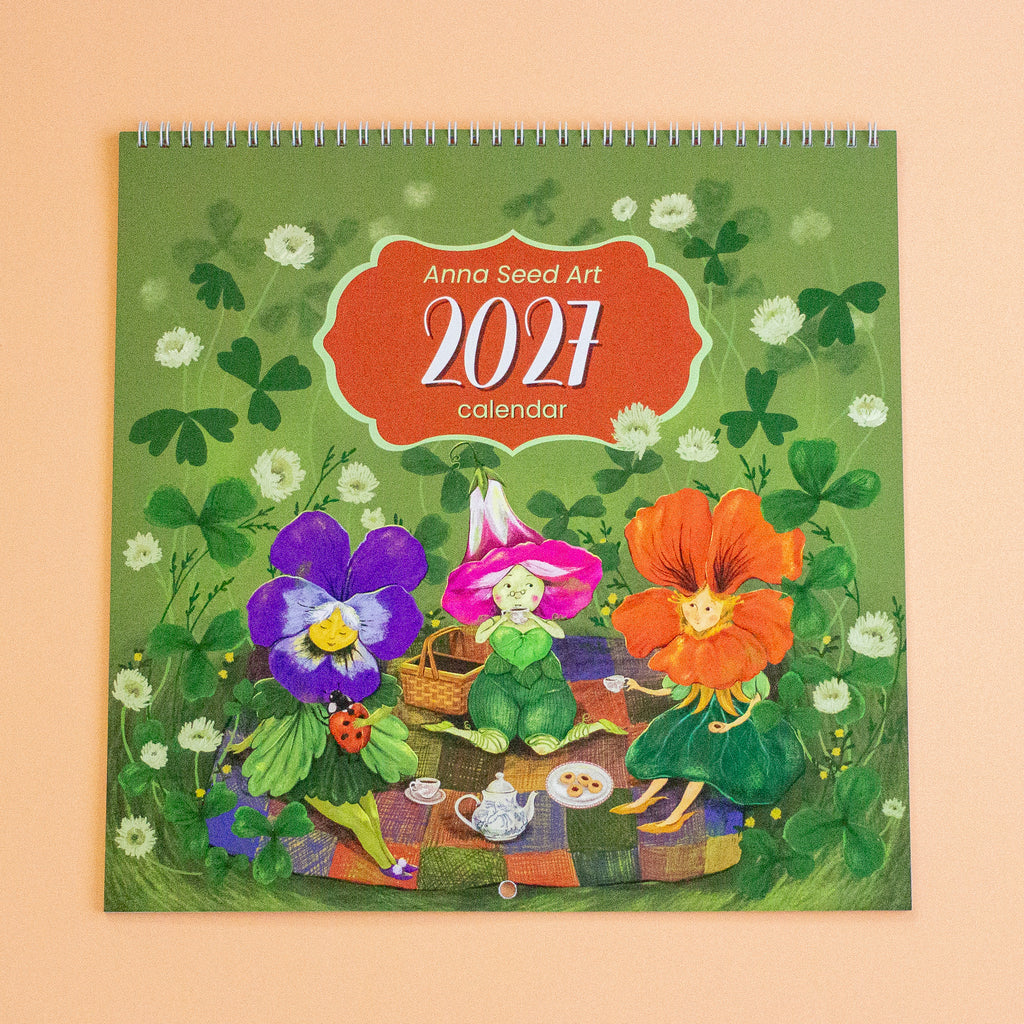 A 2027 wall calendar titled 'Anna Seed Art 2027 calendar' featuring a whimsical illustration of three flower children having a picnic on a clover field. The calendar is displayed flat against a solid peach-coloured background.