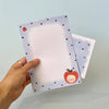 A hand holds a pastel paper with a light blue border, small dark polka dots, and a cute red apple illustration with a smiling face. Behind is the matching envelope.