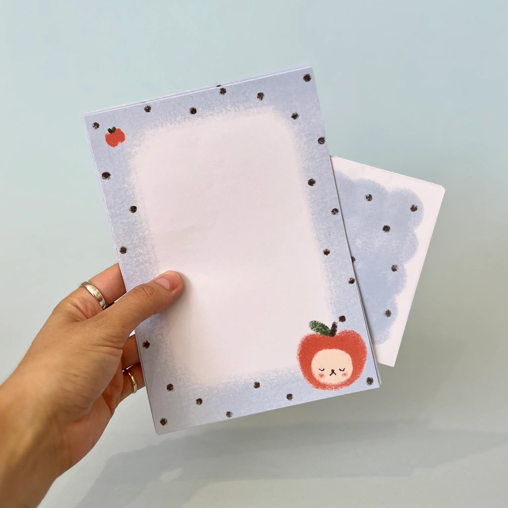 A hand holds a pastel paper with a light blue border, small dark polka dots, and a cute red apple illustration with a smiling face. Behind is the matching envelope.