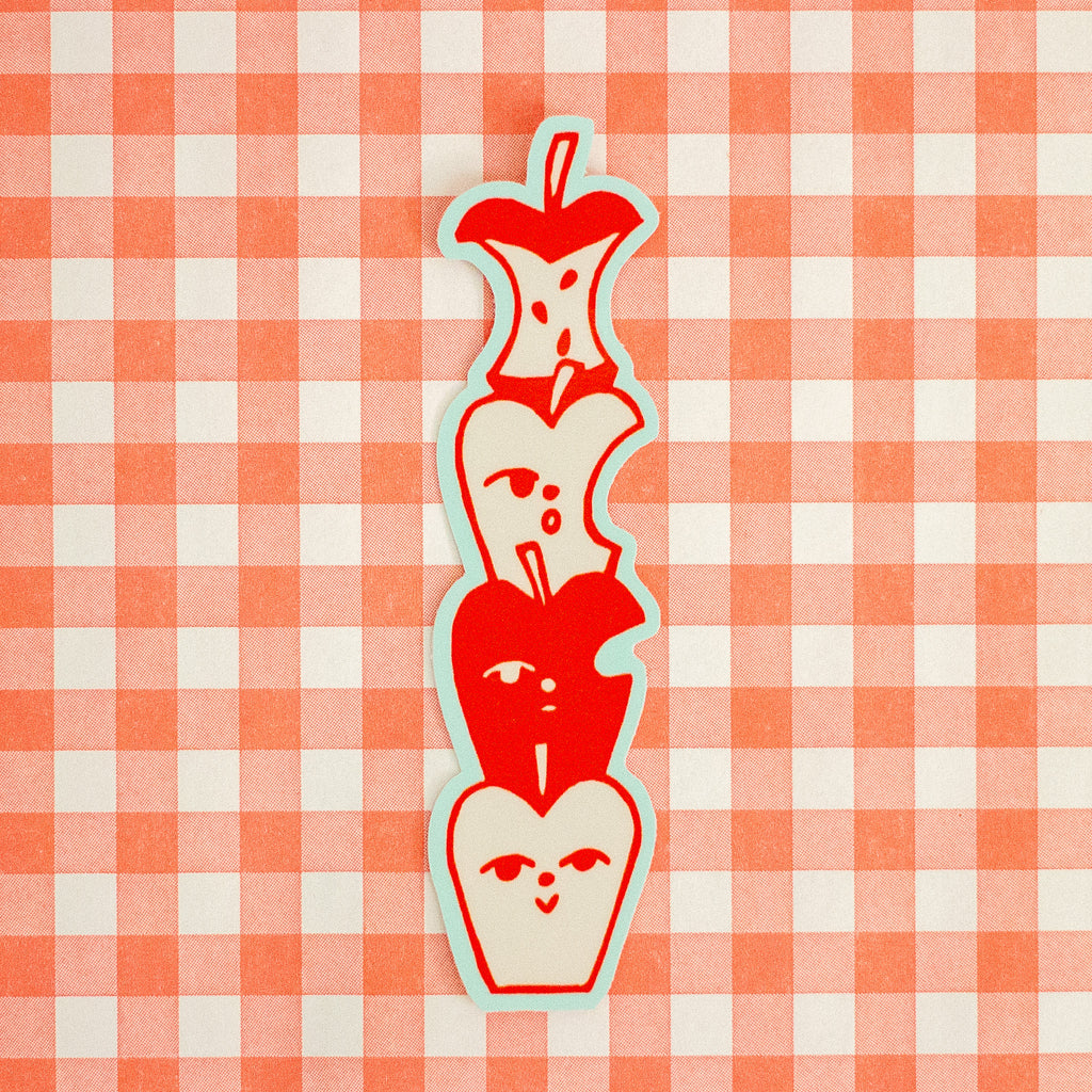 A vertical die-cut sticker featuring a stack of stylised red apple halves with simple faces, outlined in light blue, placed on a red and white gingham checkered background.