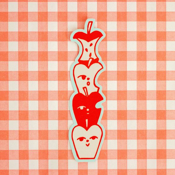 A vertical die-cut sticker featuring a stack of stylised red apple halves with simple faces, outlined in light blue, placed on a red and white gingham checkered background.