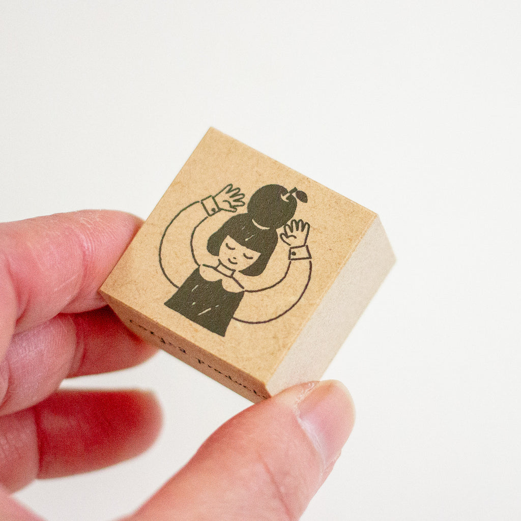 A hand holding a small square wooden stamp block with a black line drawing of a girl with an apple on her head, against a plain white background.
