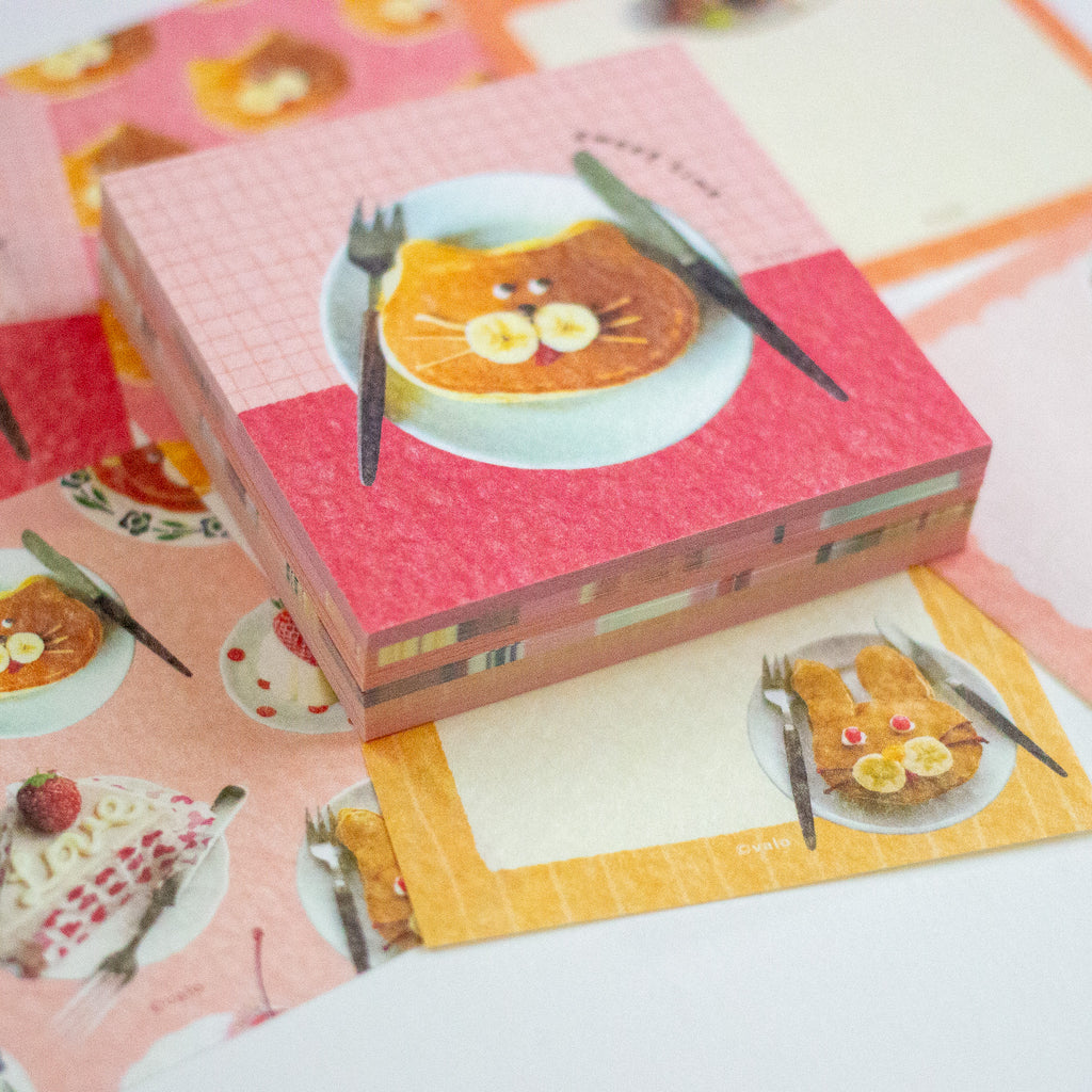 A stack of cute, pastel-coloured notepad sheets featuring a whimsical cat-shaped pancake illustration on a pink and coral background, styled on top of other matching breakfast-themed notepads.