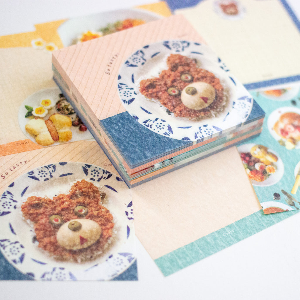 A stack of decorative paper memo sheets featuring cute food illustrations, including a bear-shaped pastry on a plate, arranged on a light surface.