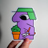 A hand holds a die-cut sticker featuring a stylised purple character with a green hat, holding orange pruning shears next to a potted plant, against a plain light background.