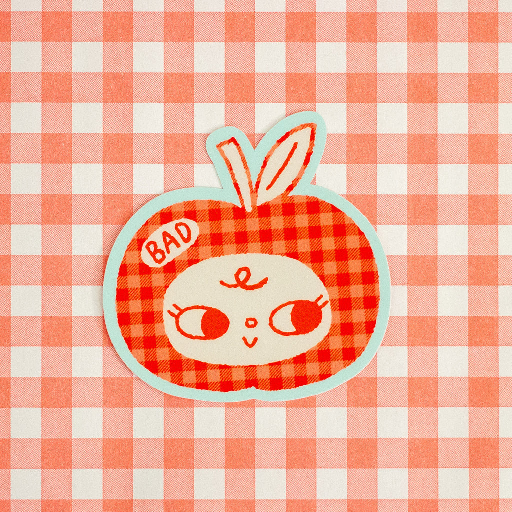 A cute, stylised apple-shaped patch or sticker featuring a red and orange gingham pattern, a white face with large eyes, and a mint green outline. The patch has a small white tag on the left that says 'BAD' in red letters.