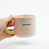 Hand holding a pink ceramic mug with a face design on a white background