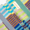 A close-up of folded striped towels in pastel and bright colours, including blue, green, pink, orange, and purple, with a small white label reading 'Odds & Ends' visible near the top.