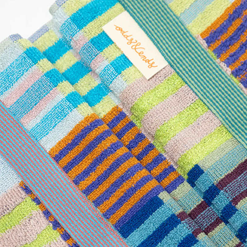 A close-up of folded striped towels in pastel and bright colours, including blue, green, pink, orange, and purple, with a small white label reading 'Odds & Ends' visible near the top.