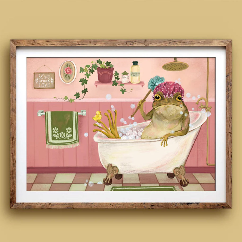 Framed artwork of a frog taking a bath in a pink bathroom setting.