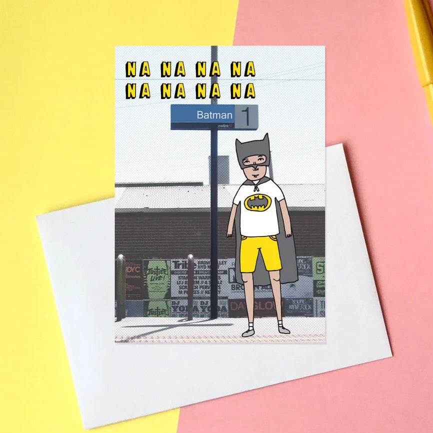 A rectangular greeting card featuring an illustrated Batman standing on a city street, placed on top of a white envelope. The card is photographed in a styled flat-lay setup on a split yellow and pink background, with a yellow pen at the top right.