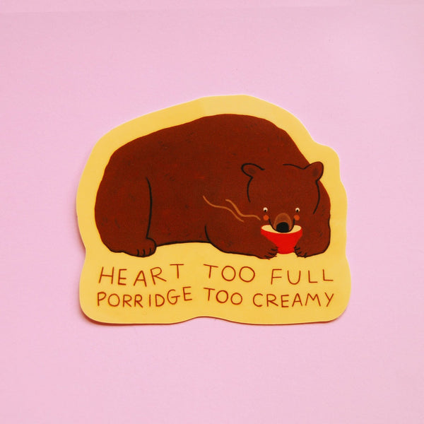 A die-cut sticker featuring a brown bear eating from a bowl with words 'HEART TOO FULL PORRIDGE TOO CREAMY' on a yellow background, set against a pink backdrop.