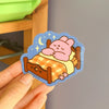 Hand holding a sticker of a bunny cartoon sleeping on a bed tucked in with a check-pattern blanket. 