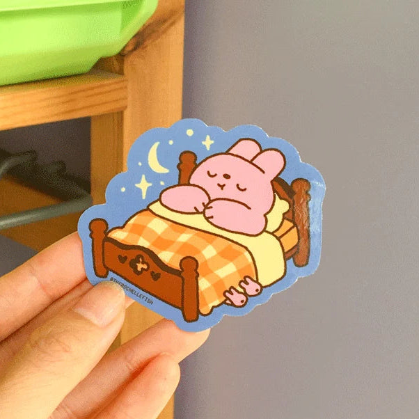Hand holding a sticker of a bunny cartoon sleeping on a bed tucked in with a check-pattern blanket. 