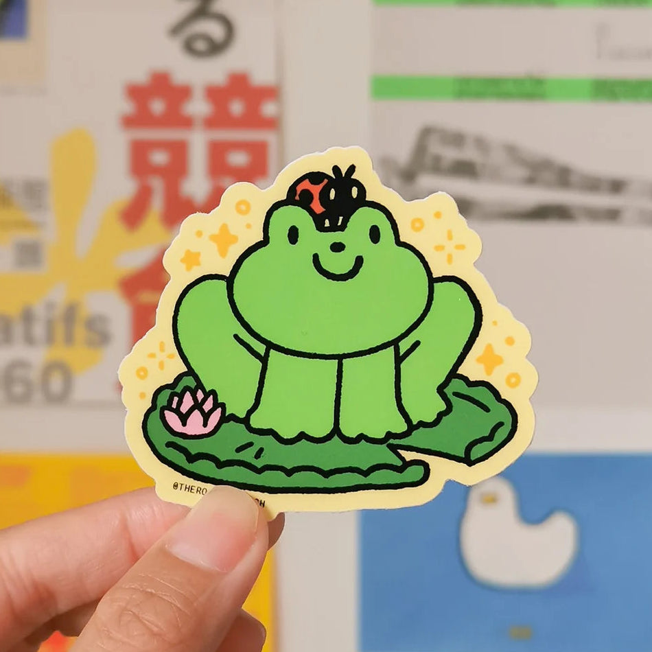 A hand holding a die-cut sticker featuring a cute cartoon green frog sitting on a lily pad with a pink lotus flower and a ladybug on its head, set against a yellow starry background. 