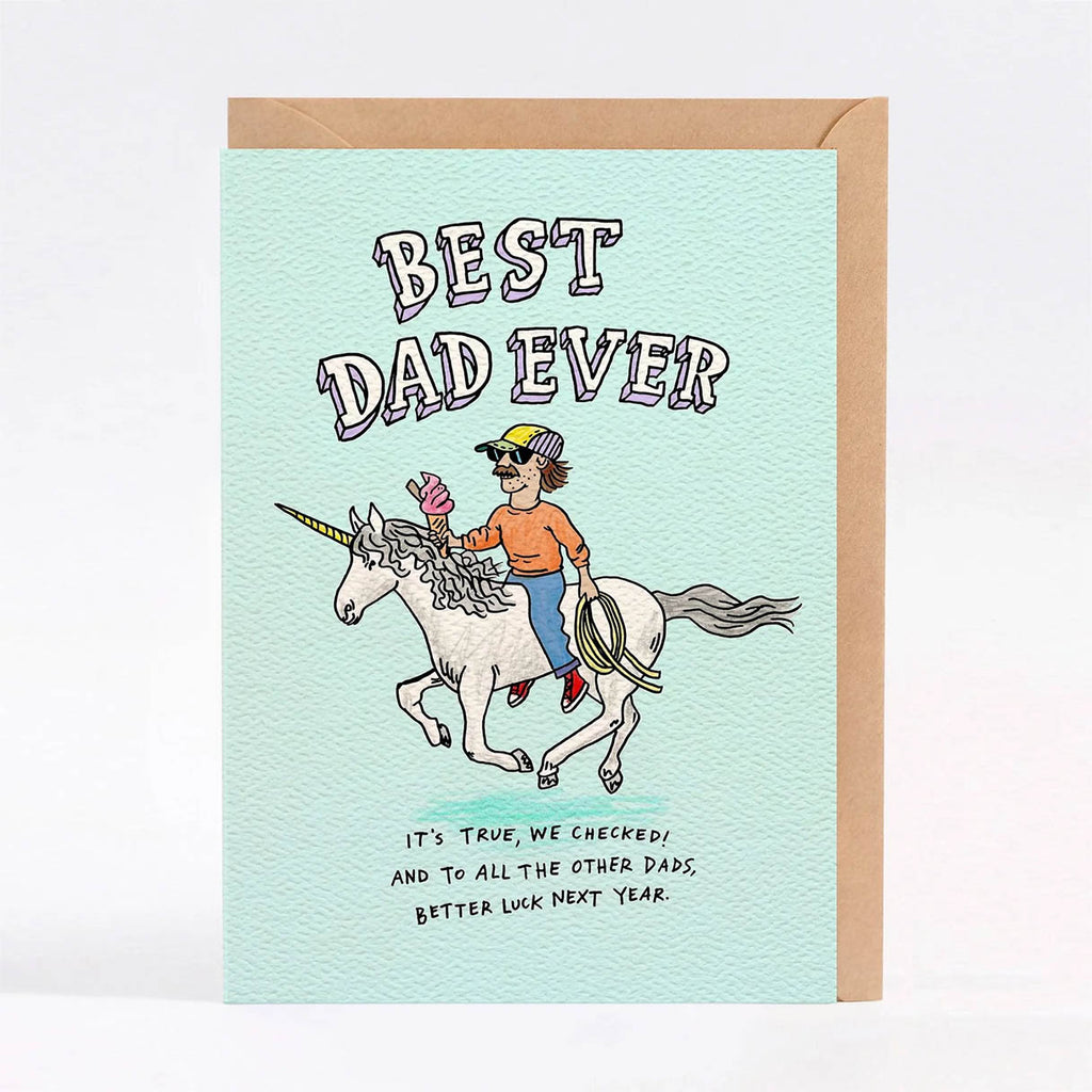 A 'Best Dad Ever' greeting card with a light mint green textured cover, featuring a cartoon illustration of a man riding a white unicorn while holding a lasso. The card is shown standing upright with a brown kraft envelope behind it against a clean white background.