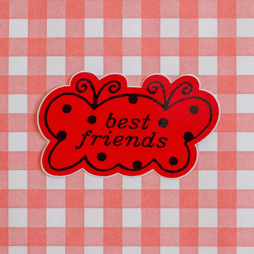 A red butterfly-shaped sticker with black polka dots and the words 'best friends' in black script, placed on a red and white checkered fabric background.