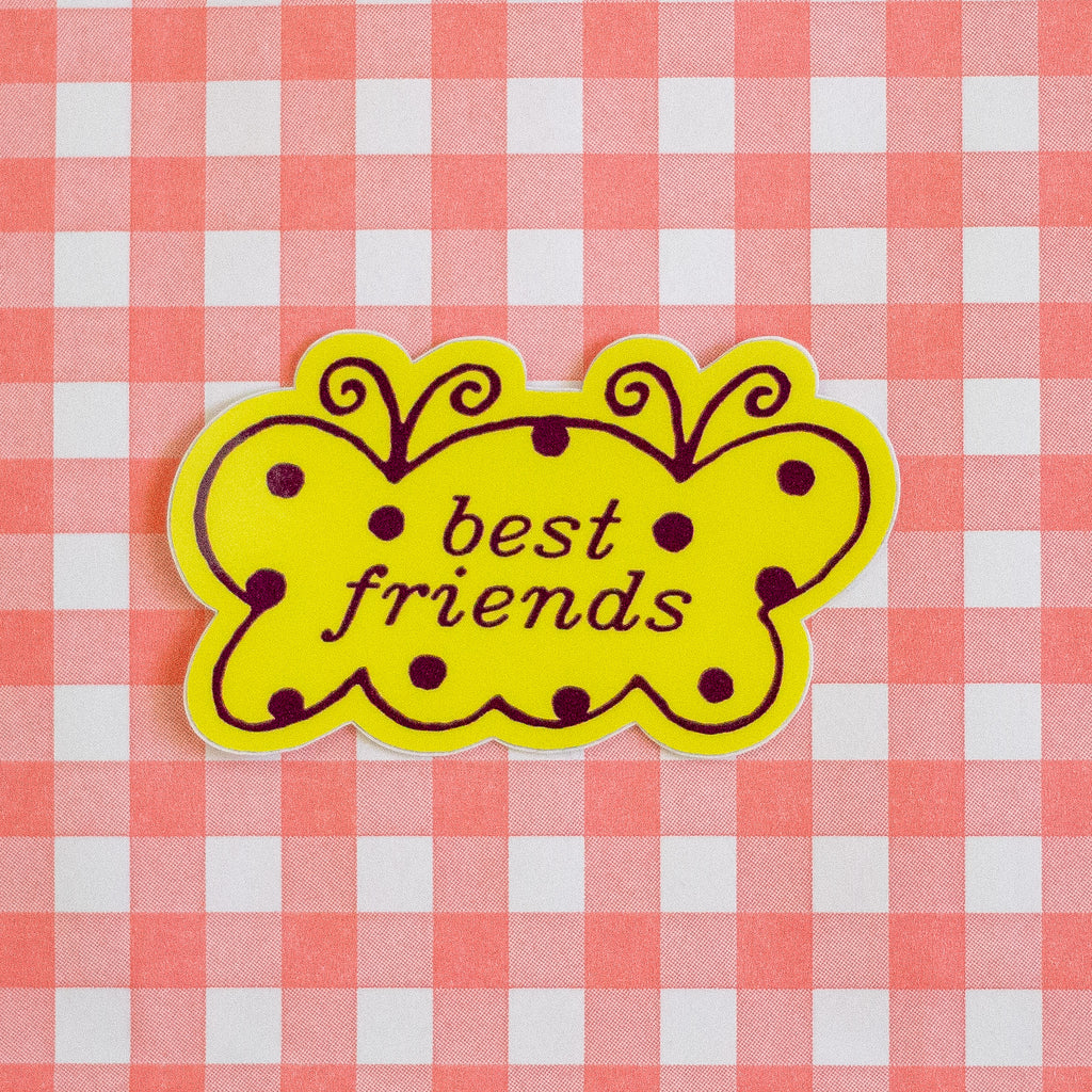 A yellow butterfly-shaped sticker with brown polka dots and the words 'best friends' in black script, placed on a red and white checkered fabric background.