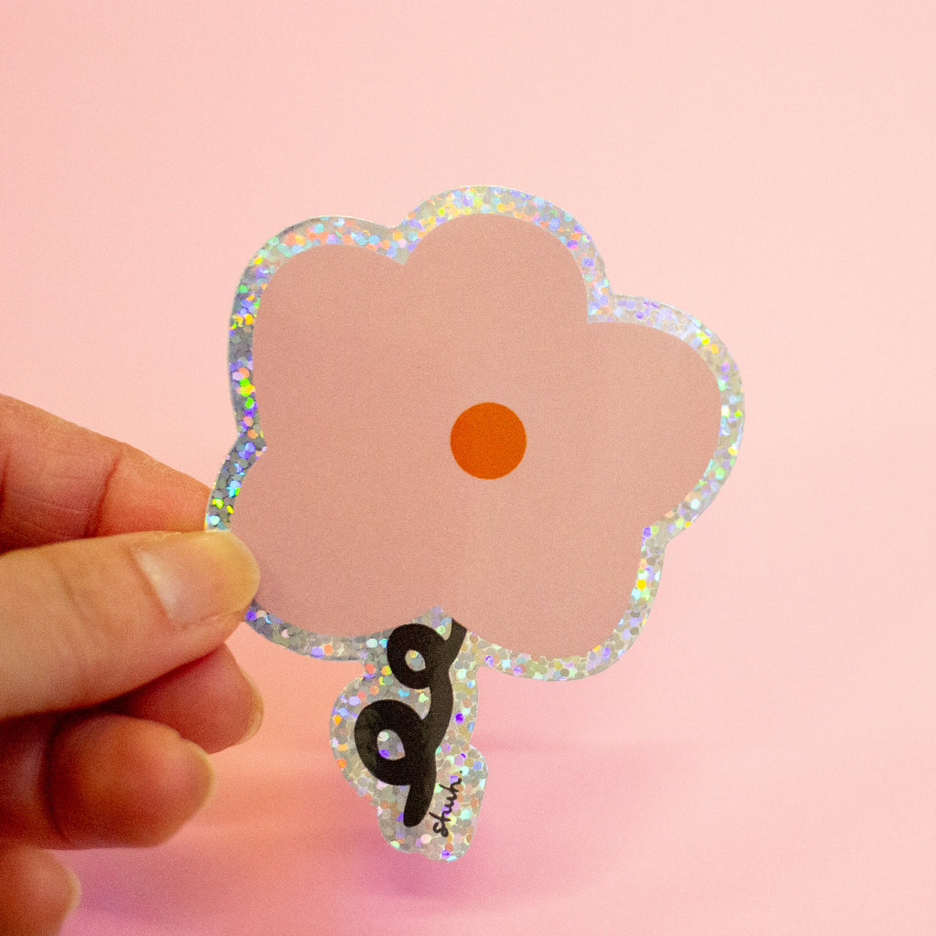 A hand holding a flower-shaped sticker with a holographic glitter border, a pale pink center, an orange circle in the middle, and a black ribbon-like accent at the bottom, set against a solid pink background.