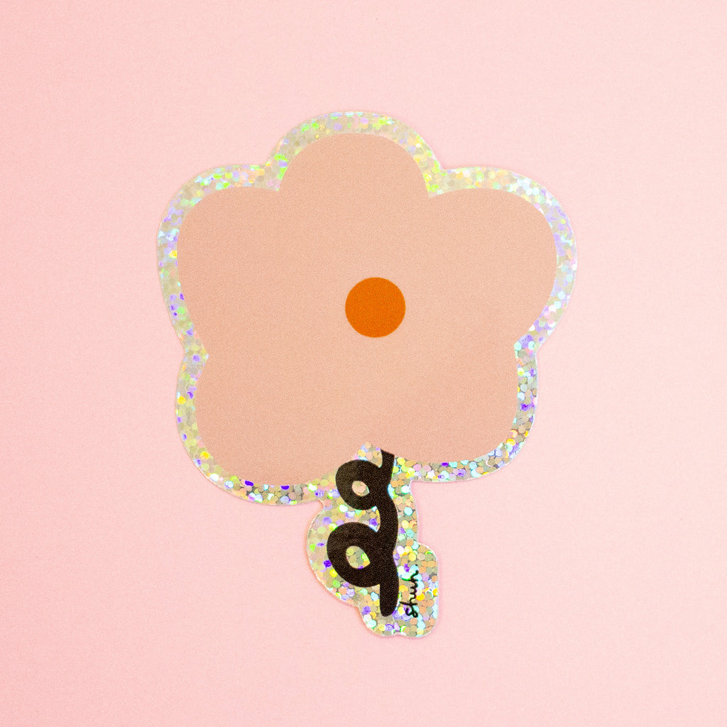 A flower-shaped die-cut sticker with a peach-pink center, a black cursive 'a' on the stem, and a holographic glitter border, set against a solid light pink background.