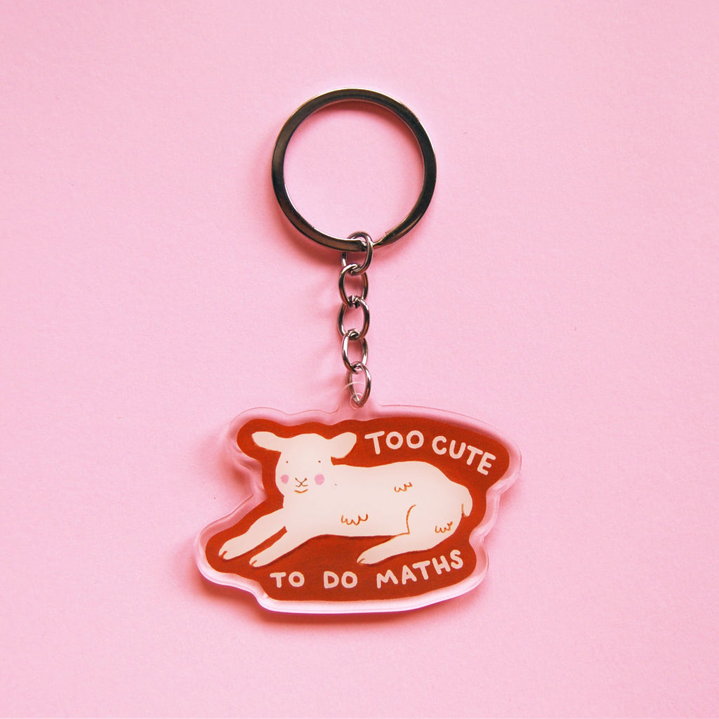 A keychain with an illustration of a lamb lying down with the words 'TOO CUTE TO DO MATHS' with a clear outline against a pink background. 
