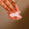 A hand holding a keychain with an illustration of a lamb lying down with the words 'TOO CUTE TO DO MATHS' with a clear outline.