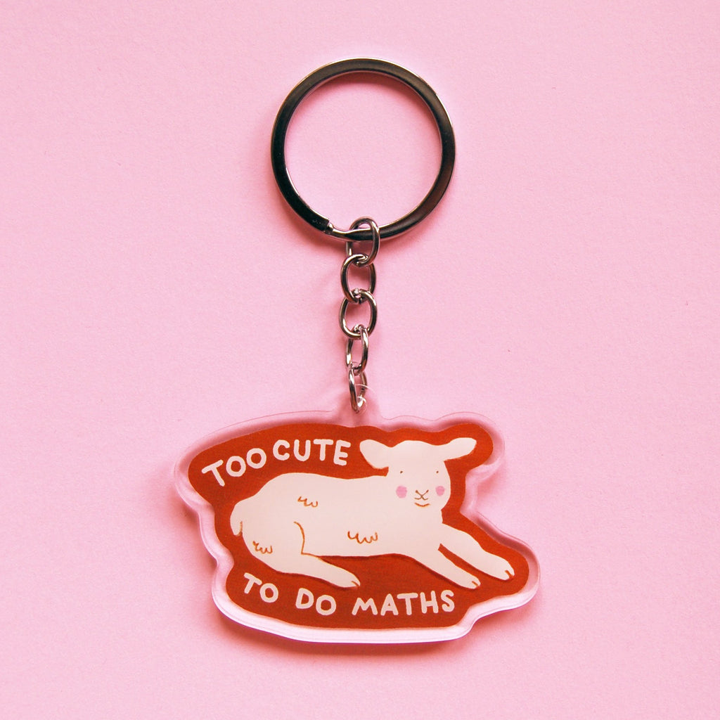 A keychain with an illustration of a lamb lying down with the words 'TOO CUTE TO DO MATHS' with a clear outline against a pink background. 