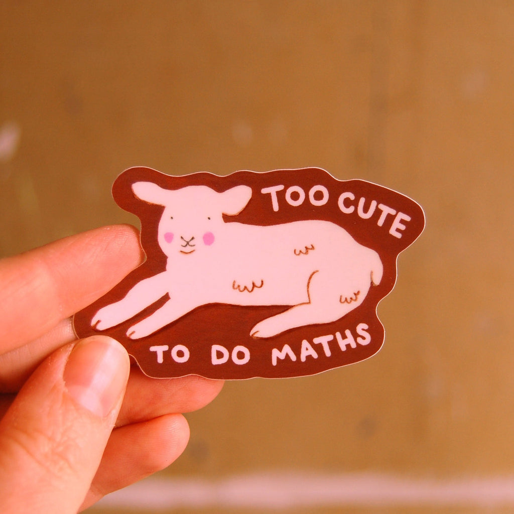 A hand holding an illustrated sticker of a lamb lying down with the words 'TOO CUTE TO DO MATHS' with a brown outline. 