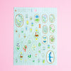 A sheet of decorative stickers featuring various floral, bird, and nature motifs on a pale blue striped background, displayed against a solid pink backdrop.
