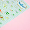 A close-up of a decorative sticker sheet featuring various floral and bird motifs on a pale mint background, placed diagonally on a solid pink surface.