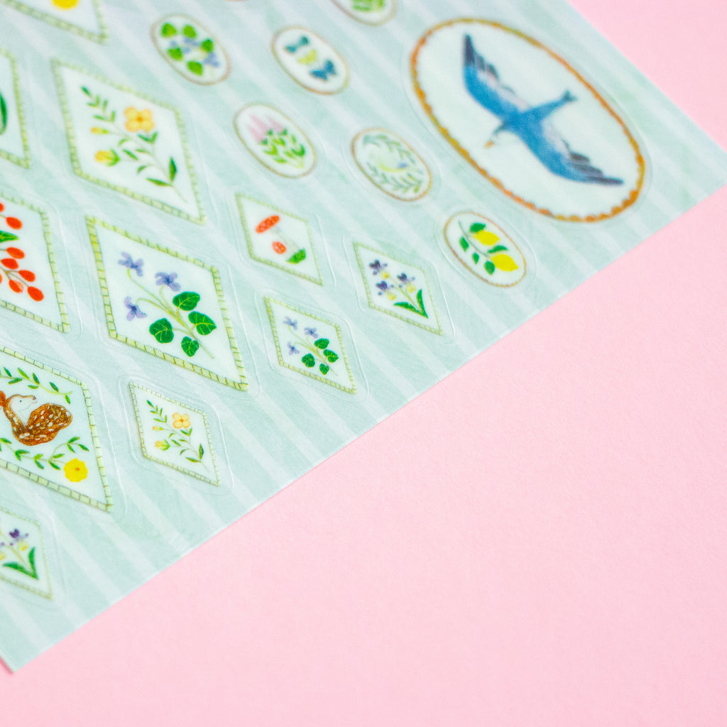 A close-up of a decorative sticker sheet featuring various floral and bird motifs on a pale mint background, placed diagonally on a solid pink surface.