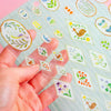 A hand holds a transparent sheet of small illustrated floral and bird stickers over a striped backing sheet on a pale pink background.
