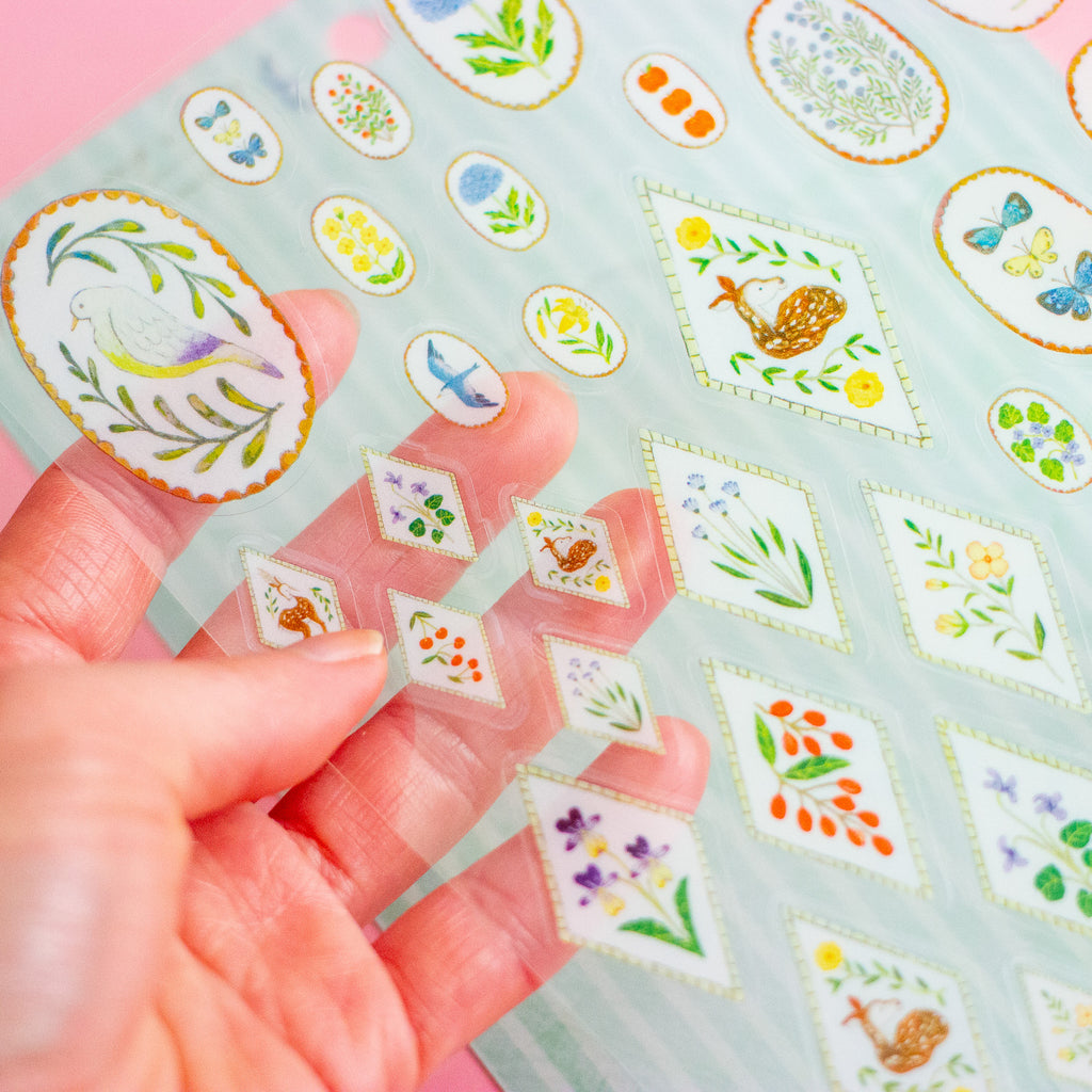 A hand holds a transparent sheet of small illustrated floral and bird stickers over a striped backing sheet on a pale pink background.