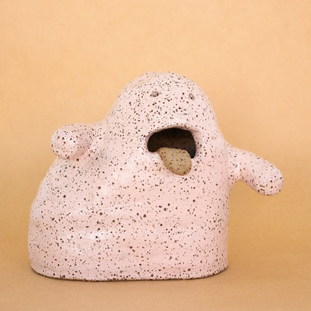 A front view of the Handbuilt ceramic figurine by shuh. with a speckled pink glaze, open mouth, and protruding tongue. Sculptural artwork made from stoneware clay for the Well Wishes exhibition.