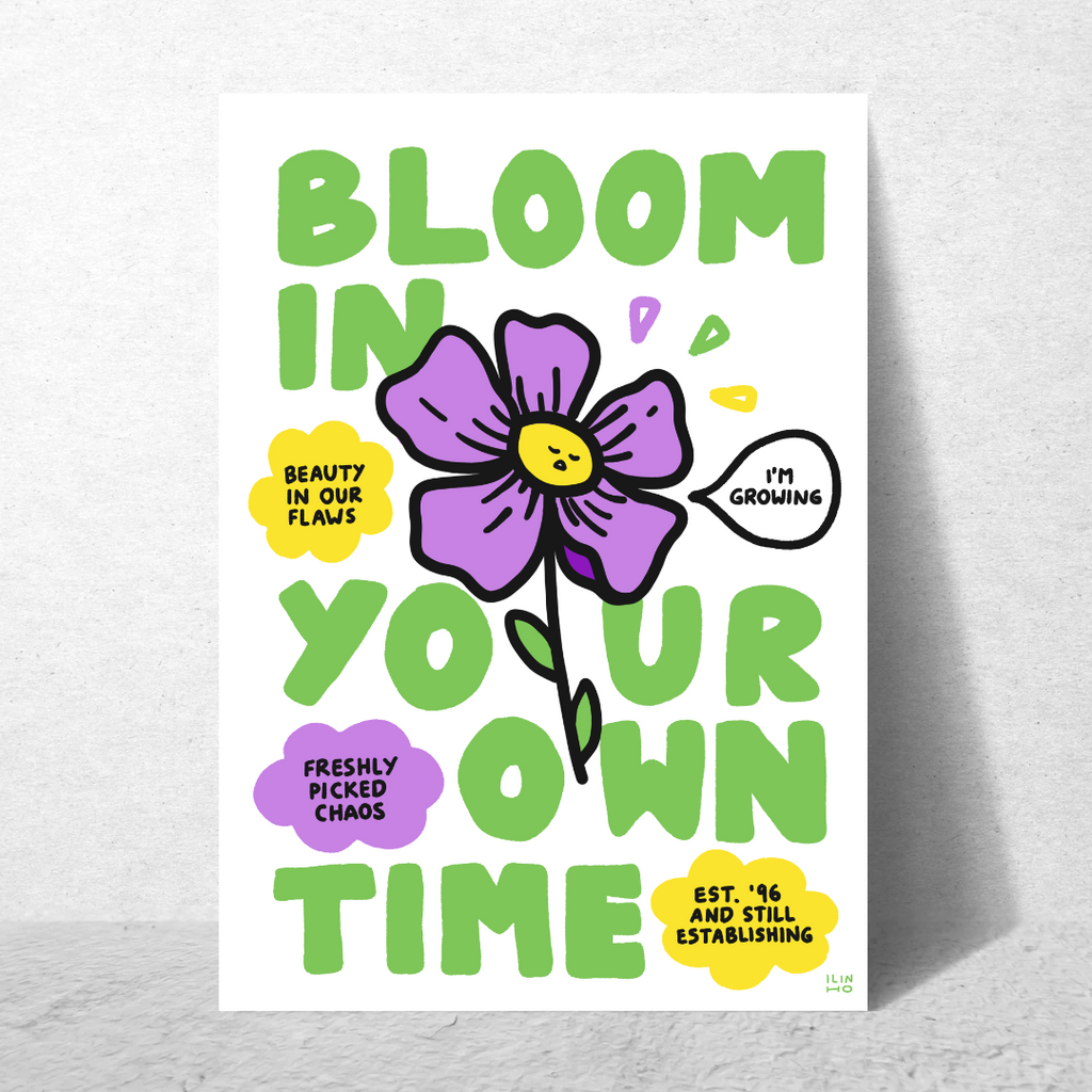 A vertical art print with a white background featuring a large purple flower and bold green text that reads 'BLOOM IN YOUR OWN TIME,' with smaller decorative text bubbles in yellow and purple, shown leaning against a light grey wall.