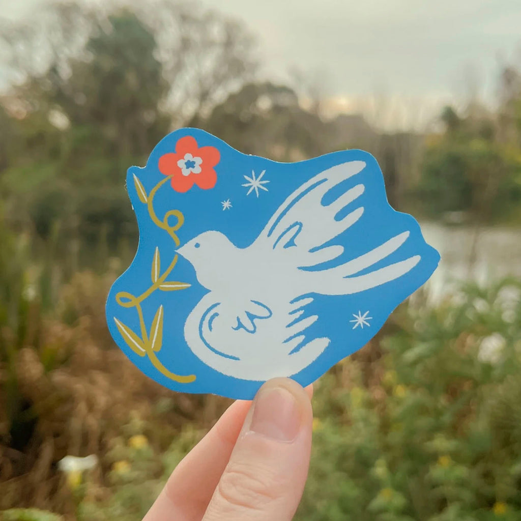 A hand holds a blue die-cut sticker featuring a white dove, a red flower, and yellow leaves against an outdoor blurred background.