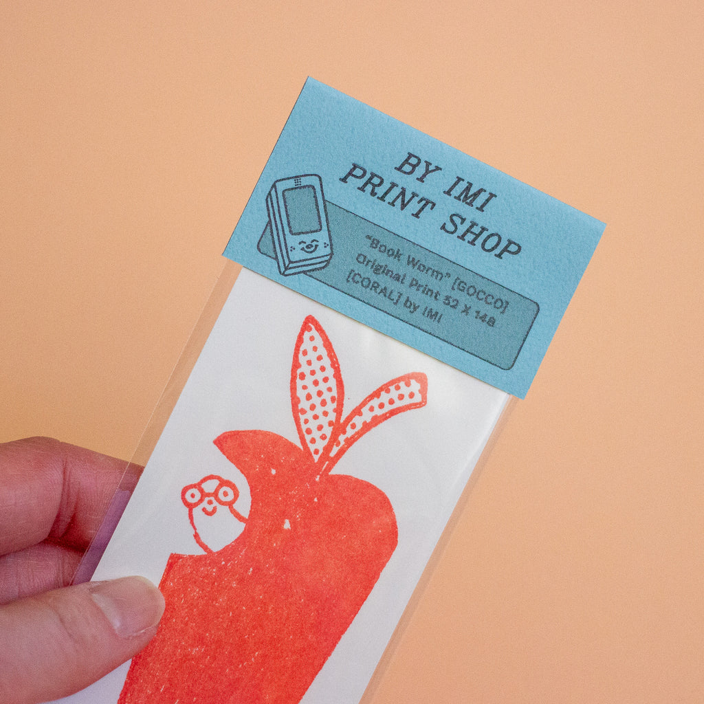 A hand holds a printed card package featuring a red apple illustration on a white background, with a light blue header card that reads 'BY IMI PRINT SHOP' and 'Book Worm [GOCCO] Original Print 52 x 148 [CORAL] by IMI.'.