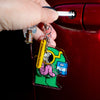 A hand inserts a key into a car door lock while holding a colourful cartoon keychain charm. The charm features a green character with pink and blue details, attached to a yellow tag that reads 'Samantha Curcio'.
