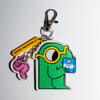A cartoon keychain featuring a green cartoon figure with yellow glasses holding a blue book, attached to a silver lobster clasp. The keychain also includes a pink worm charm and a yellow tag printed with 'Samantha Curcio'.