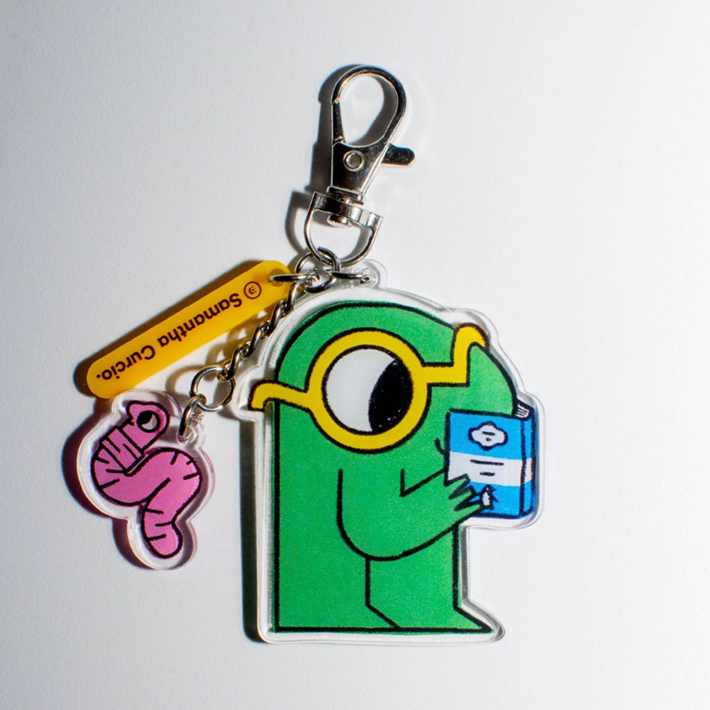 A cartoon keychain featuring a green cartoon figure with yellow glasses holding a blue book, attached to a silver lobster clasp. The keychain also includes a pink worm charm and a yellow tag printed with 'Samantha Curcio'.