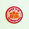 A circular embroidered patch with a red border and the text 'BORN TO BE AN ADVENTURER' surrounding a smiling orange and yellow butterfly on a pale mint background.