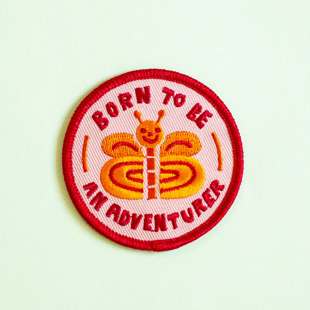 A circular embroidered patch with a red border and the text 'BORN TO BE AN ADVENTURER' surrounding a smiling orange and yellow butterfly on a pale mint background.