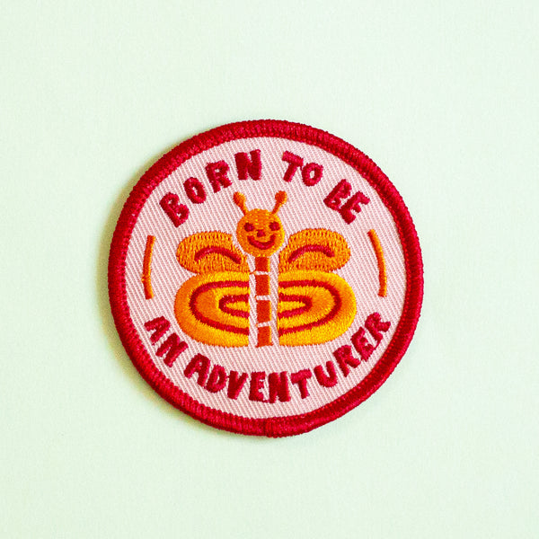 A circular embroidered patch with a red border and the text 'BORN TO BE AN ADVENTURER' surrounding a smiling orange and yellow butterfly on a pale mint background.