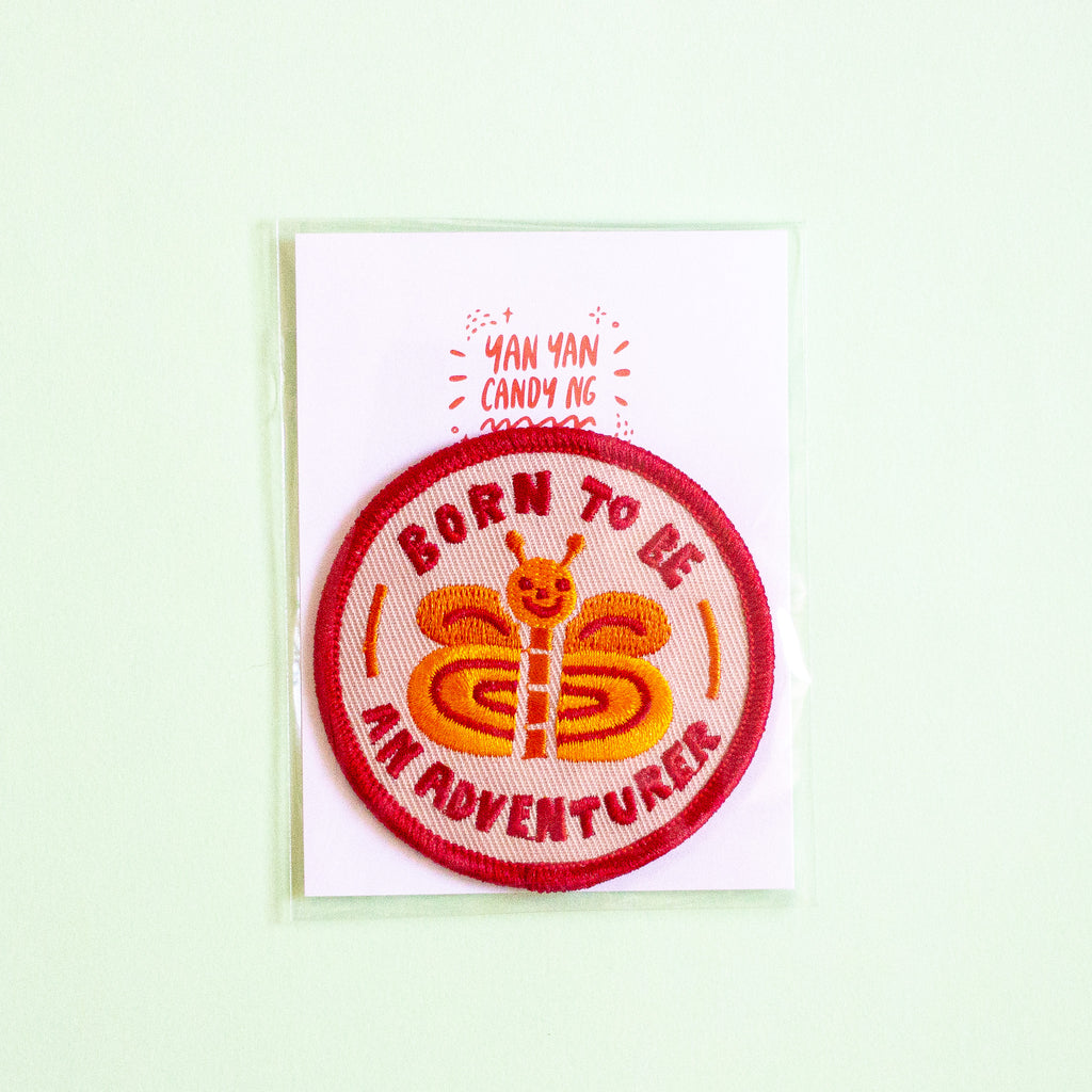 A circular embroidered patch featuring a smiling orange and yellow butterfly with the text 'BORN TO BE AN ADVENTURER' in red, displayed on a white backing card that reads 'YAN YAN CANDY NG'. The card is photographed against a pale mint green background.