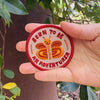 A hand holding a round embroidered patch that reads 'BORN TO BE AN ADVENTURER' with a butterfly design, set against a background of green leaves and a brown tree trunk.