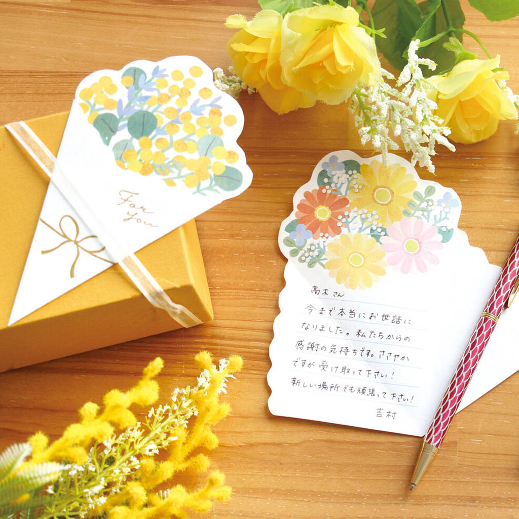 Two decorative Japanese-style note cards with floral illustrations are arranged on a wooden surface with yellow roses, a gold gift box, and a red-and-white striped pen. One card is shown folded over the box with the words 'For you,' and the other is open with handwritten text.